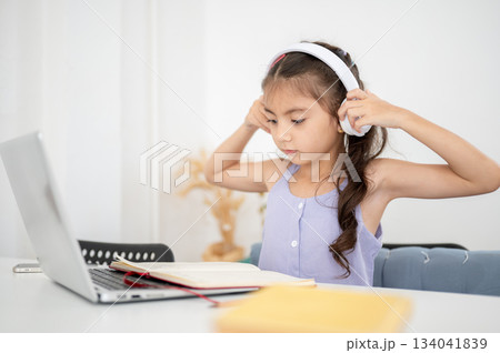 Asian kid girl putting on or taking off headphones while sitting with laptop at white table in house Asian kid girl putting on or taking off headphones while sitting with laptop at white table in house 134041839