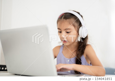 Asian kid or girl with headphones looking at laptop with open mouth while sitting at table in house. 134041851