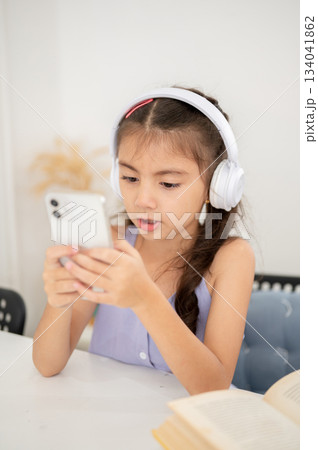 Asian kid or girl with headphones looking at phone in hands with mouth open while sitting at table. Asian kid or girl with headphones looking at phone in hands with mouth open while sitting at table. 134041862