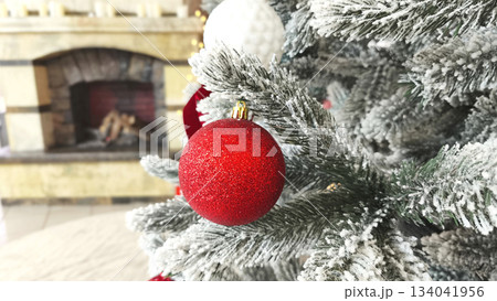 A close-up of a red Christmas bauble on a snow-covered tree branch with a blurred fireplace in the background. A warm and festive atmosphere for seasonal designs and winter themes. A close-up of a red Christmas bauble on a snow-covered tree branch with a blurred fireplace in the background. A warm and festive atmosphere for seasonal designs and winter themes. 134041956