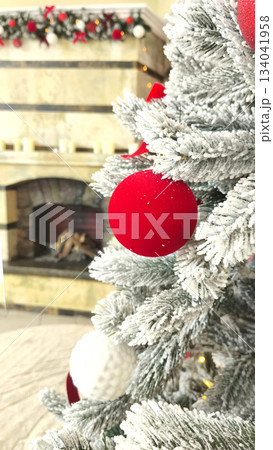 A vertical close-up of a red Christmas ball on a snow-covered Christmas tree branch with a blurred fireplace in the background. A festive winter interior decor element. A vertical close-up of a red Christmas ball on a snow-covered Christmas tree branch with a blurred fireplace in the background. A festive winter interior decor element. 134041958