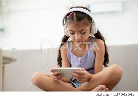 Asian kid or girl with headphones is looking at her phone while sitting cross legged on fabric sofa. 134042043