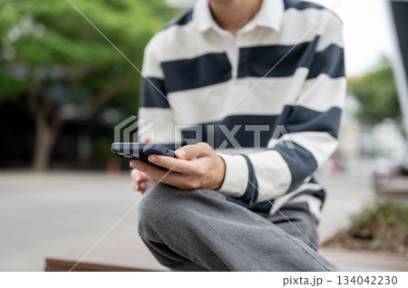 Close up of a man holding smartphone while sitting cross legged on a wooden bench in the parking lot Close up of a man holding smartphone while sitting cross legged on a wooden bench in the parking lot 134042230