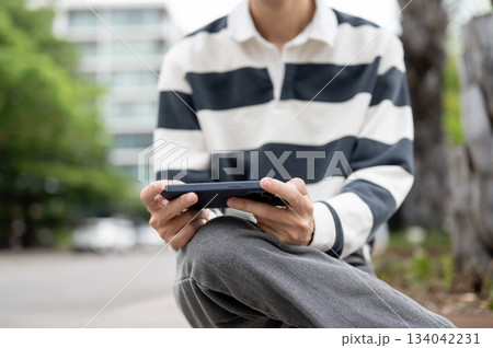 Close up of man holding phone horizontally while sitting cross legged on wooden bench in parking lot 134042231