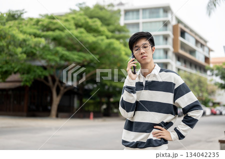 Asian man wearing glasses having a phone call with a hand on his waist while standing in parking lot Asian man wearing glasses having a phone call with a hand on his waist while standing in parking lot 134042235