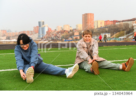 Two Russian women aged 50 and 60 stretch forward while sitting on grass at a stadium in Vladivostok, staying active together and supporting healthy life 134043964