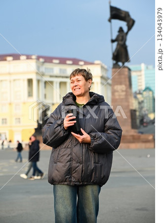 Russian woman 60 years old stands on city square in Vladivostok holding a thermal mug of coffee enjoying calm moment and morning mindfulness and peace 134043979