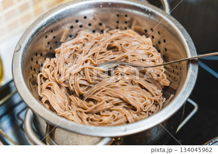 Pasta, buckwheat noodles cooking in a colander on the stove in a kitchen setting with a fork Pasta, buckwheat noodles cooking in a colander on the stove in a kitchen setting with a fork 134045962