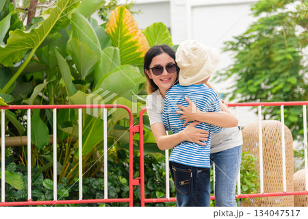 Smiling mother woman embraces child son, sharing warm family bond during happy summer city day, parent people enjoy outdoor urban street activity amidst lush green foliage. Smiling mother woman embraces child son, sharing warm family bond during happy summer city day, parent people enjoy outdoor urban street activity amidst lush green foliage. 134047517
