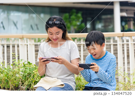 Mother, child, summer city scene, family woman and son enjoy playing mobile games outdoors, parent and child connecting with technology on urban street, people having fun. Mother, child, summer city scene, family woman and son enjoy playing mobile games outdoors, parent and child connecting with technology on urban street, people having fun. 134047520