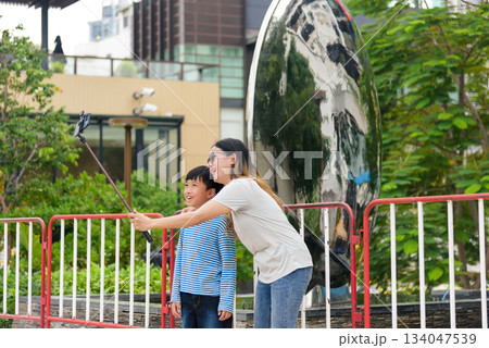 Mother, son, child, family, woman, parent, people find summer joy on city street, taking cheerful selfie outdoors, urban background shows vibrant foliage, reflective buildings. 134047539