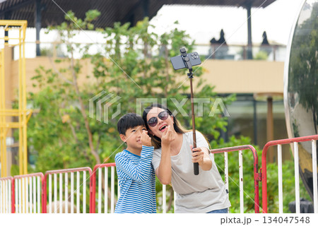 Mother, child enjoy summer city street, happy family woman parent son taking selfie with phone on selfie stick, making finger heart sign, playful bonding outdoors, green trees urban setting. 134047548