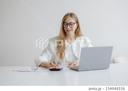 Young woman working on laptop with calculator during daytime in a minimalistic workspace 134048316