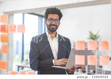 An Indian businessman smiles while holding a laptop in a modern office setting 134048584