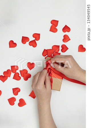 Hands Tie Red Ribbon Around Box on White Table With Heart Decorations for Valentine's Day 134049545