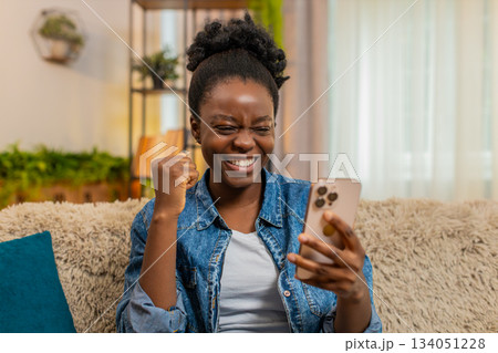 African American young woman celebrates exam results and game victory with smartphone at home African American young woman celebrates exam results and game victory with smartphone at home 134051228