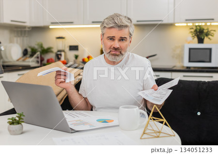 Middle-aged man puzzled by documents invoices at table with laptop shocked by very big costs at home 134051233