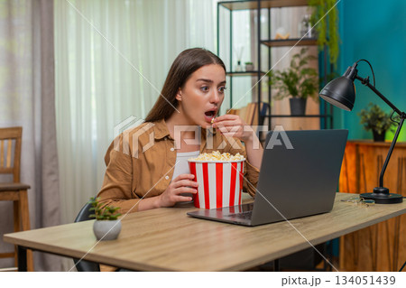 Young woman watching dramatic movie on laptop eating popcorn nervously reacting to intense scene Young woman watching dramatic movie on laptop eating popcorn nervously reacting to intense scene 134051439