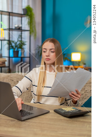 Caucasian woman managing tax declaration at home on sofa table, holding documents, calculator laptop 134052244