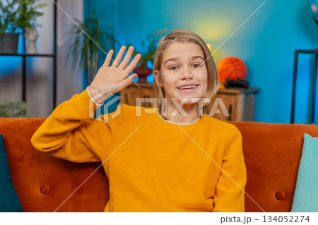 Teenager boy waving with smile greeting happily sitting on sofa, looking straight at camera at home Teenager boy waving with smile greeting happily sitting on sofa, looking straight at camera at home 134052274