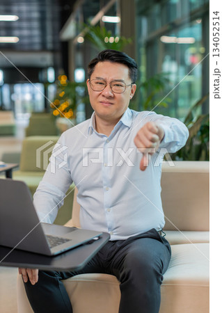 Upset businessman in shirt with laptop computer showing thumb down gesture in modern office lobby Upset businessman in shirt with laptop computer showing thumb down gesture in modern office lobby 134052514
