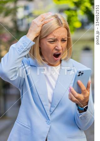 Caucasian mature businesswoman looking shocked while reading bad news on mobile phone on city street 134052551