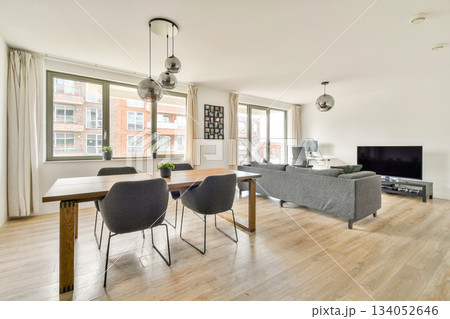 A stylish and contemporary living room featuring a wooden dining table, comfortable seating, and large windows allowing natural light into the space. 134052646