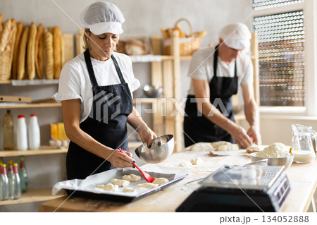 Two bakers prepare dough for baking - they put pieces of raw dough on baking sheet and pour oil over them 134052888