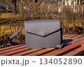 Stylish gray crossbody handbag on a wooden bench in a park during a sunny day. eco leather bag with a long strap, shoulder bag 134052890