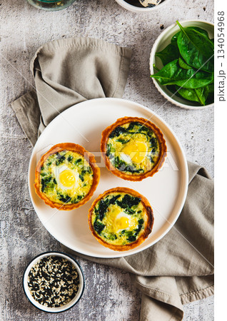 Eggs, spinach and cheese baked in tartlets on a plate on the table top and vertical view Eggs, spinach and cheese baked in tartlets on a plate on the table top and vertical view 134054968