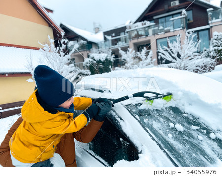 Child removes snow from a car on a winter day between houses covered in snow, with trees and other vehicles visible in the neighborhood Child removes snow from a car on a winter day between houses covered in snow, with trees and other vehicles visible in the neighborhood 134055976