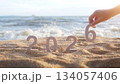 hand place the number 6 on sandy beach, transition from year 2025 to 2026 for new year sign on summer beach at sunset time, with ripple sea wave and beautiful nature at background, New Year celebrate 134057406