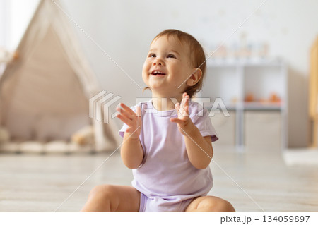 Smiling infant baby girl claps hands while sitting on floor at home Smiling infant baby girl claps hands while sitting on floor at home 134059897