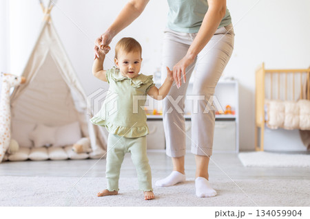 Baby practicing first steps while holding mothers hands in bright home interior Baby practicing first steps while holding mothers hands in bright home interior 134059904