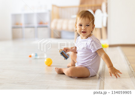 Smiling baby sitting on floor holding small toy in bright home interior Smiling baby sitting on floor holding small toy in bright home interior 134059905