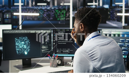 Server farm computer scientist interprets AI generated data analytics graphs. African american man in data center using artificial intelligence program showing predictive system metrics, camera A Server farm computer scientist interprets AI generated data analytics graphs. African american man in data center using artificial intelligence program showing predictive system metrics, camera A 134060209