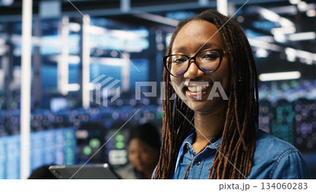 Portrait of smiling server room engineer using device to interpret AI generated data analytics graphs. Happy data center worker using artificial intelligence tool showing system metrics, camera B Portrait of smiling server room engineer using device to interpret AI generated data analytics graphs. Happy data center worker using artificial intelligence tool showing system metrics, camera B 134060283