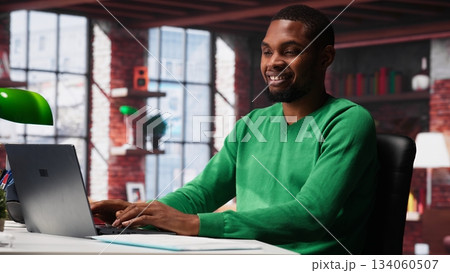 Black remote employee in home office using laptop for web assignments, preparing presentation slides and working on data layouts. Young teleworker finishing company project. Camera A. 134060507