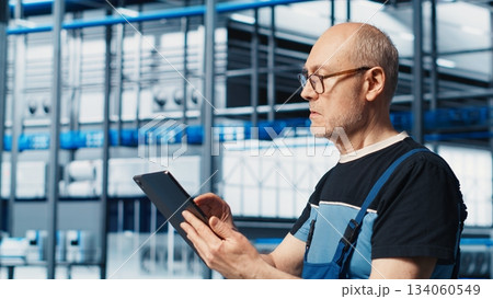 Senior engineer in smart photovoltaics factory looking at system performance metrics on tablet. Experienced solar panels plant worker tracking energy production through IoT sensors on device, camera A 134060549