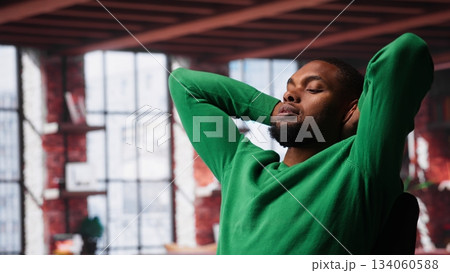 Man at home deals with back pain and stretching joints on chair, being uncomfortable due to incorrect posture. Black guy feeling overworked and experiencing muscle cramps at desk. Camera A. Man at home deals with back pain and stretching joints on chair, being uncomfortable due to incorrect posture. Black guy feeling overworked and experiencing muscle cramps at desk. Camera A. 134060588