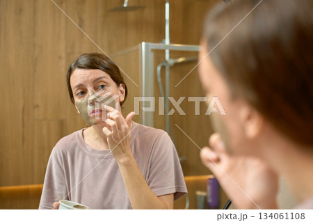 Woman applying clay face mask and looking at reflection in bathroom mirror Woman applying clay face mask and looking at reflection in bathroom mirror 134061108