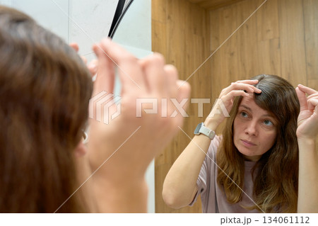 Woman looking at gray hair and scalp parting in bathroom mirror 134061112