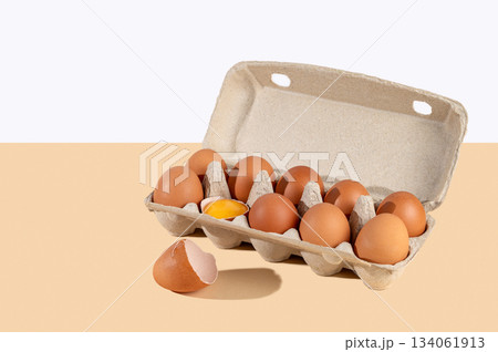 Fresh brown eggs in an open carton with one cracked yolk on a beige and white background 134061913