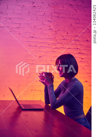 Smiling woman holding cup while working on laptop in neon workspace 134062425