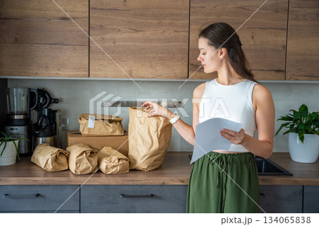 Young woman checking a list while looking at unpacked meal kit ingredients organized by recipe. Concept of planning weekly cooking using convenient pre-portioned grocery delivery. Young woman checking a list while looking at unpacked meal kit ingredients organized by recipe. Concept of planning weekly cooking using convenient pre-portioned grocery delivery. 134065838