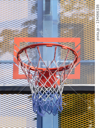 An orange metal basketball rim and net are mounted on a fiberglass backboard, prominently framed by a steel grid fence. 134066186