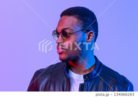 Portraif of fashionable black man in stylish leather jacket in neon light standing over purple background. Young handsome african american guy posing in studio, vertical shot, free space 134066265