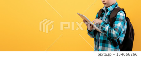 Ginger kid schooler using digital tablet for education, doing homework on yellow studio background, copy space. Smiling redhead boy with backpack holding pad, websurfing or using new application 134066266