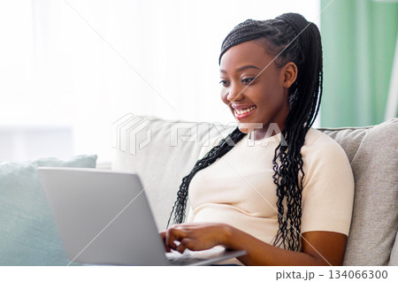 Happy young black woman with cute hair freelancer working from home, sitting on couch and using modern laptop, typing text or communicating with clients online, blogging or copywriting, copy space 134066300