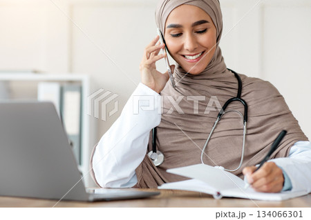 Happy muslim woman in hijab doctor taking notes while talking on mobile phone, having conversation with sick patient, giving recommendations, prescribing treatment on phone, free space 134066301